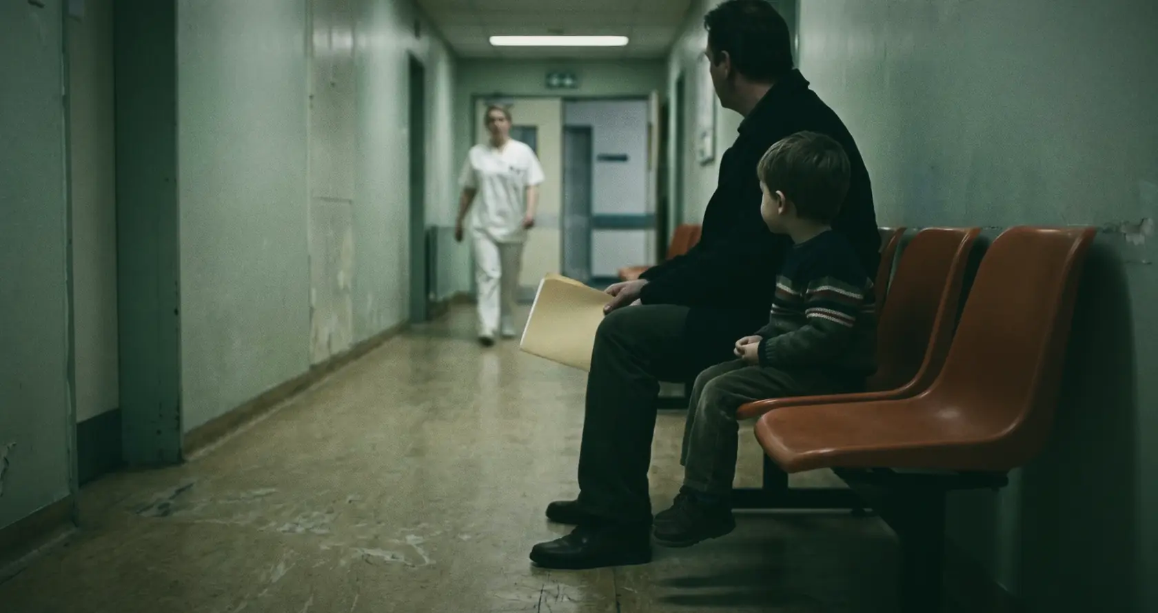 A man and a small boy, seen from behind, waiting on orange chairs in a pale green clinic corridor.