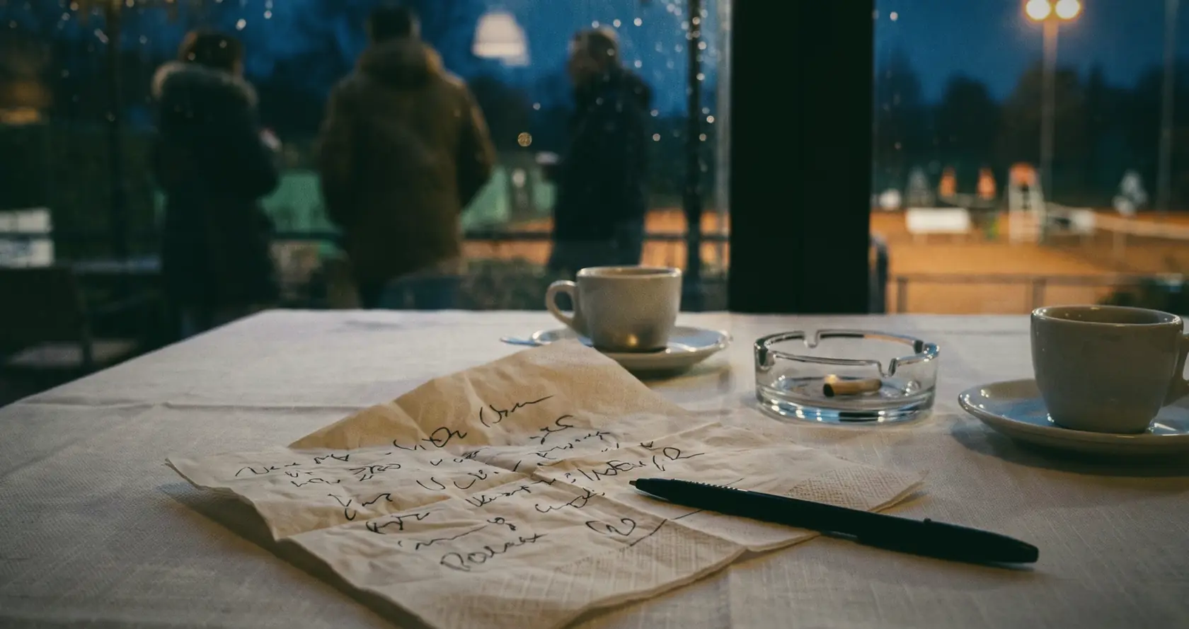 A handwritten paper napkin, pen and coffee cups on a restaurant table, by a window at dusk.