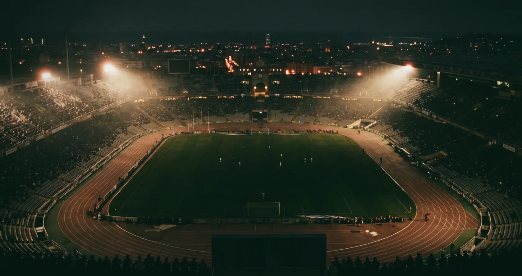 An athletics stadium at night from high above, floodlights in haze and a city behind.