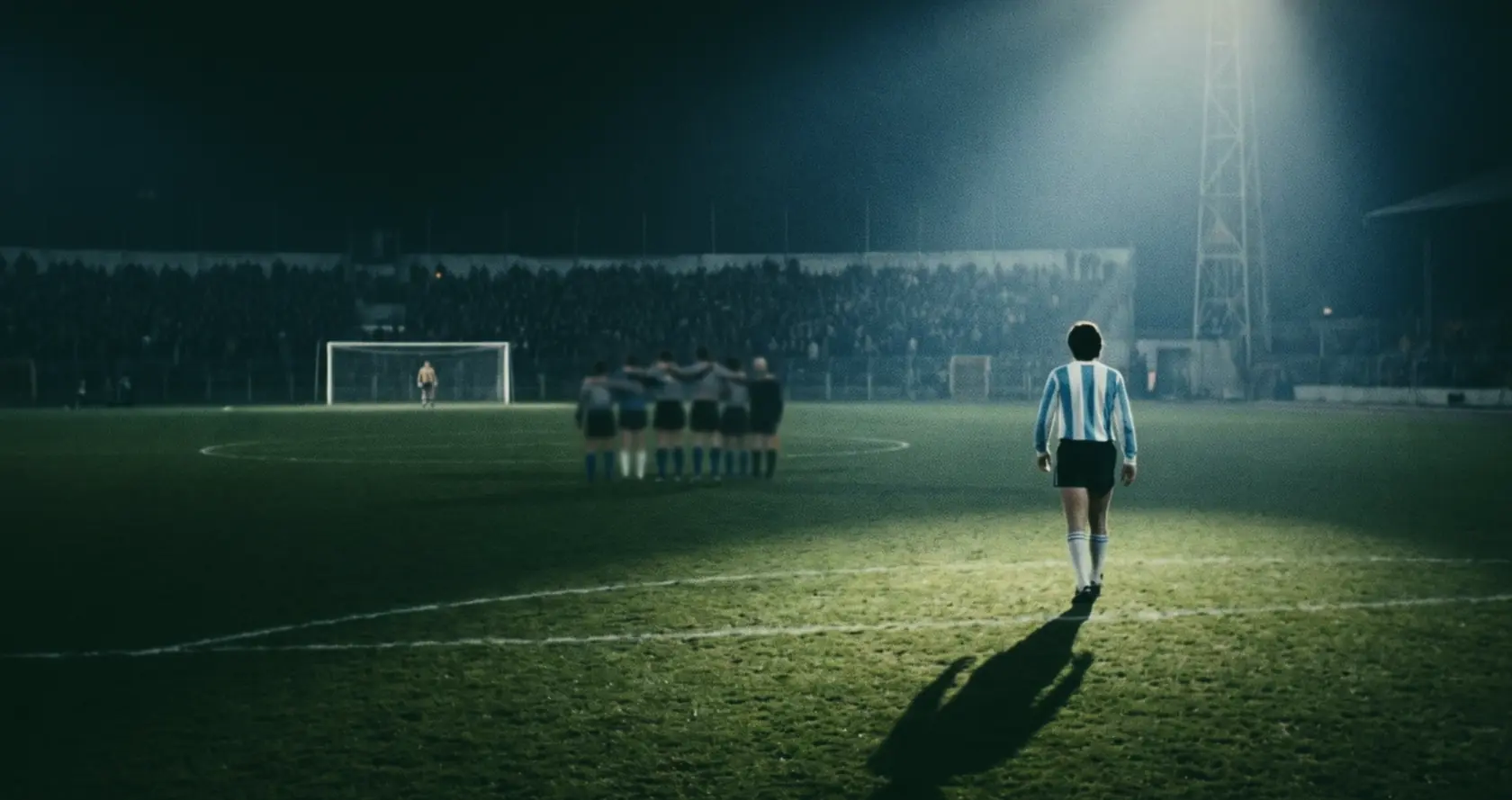 A lone player in pale blue and white stripes walking towards a distant goal and a huddled team, at night.