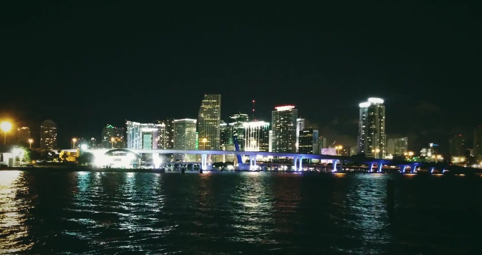 The Miami skyline at night across dark water, with coloured reflections.